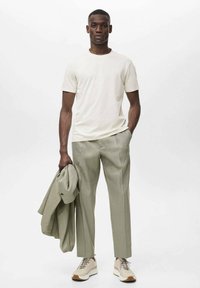 T-shirt beige chiaro, pantaloni verdi chiari su misura, giacca grigia appoggiata sul braccio e sneakers beige con accenti marroni. Design semplice e moderno.