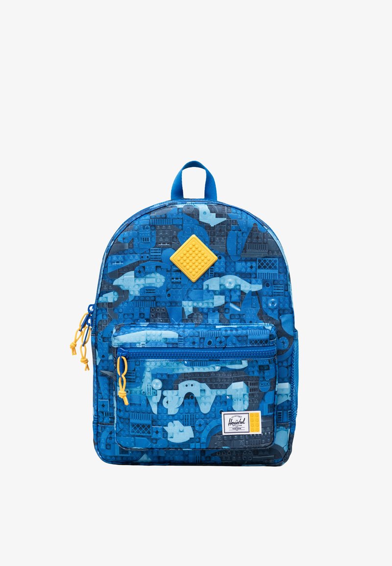 Sac à dos bleu avec motif de briques de type LEGO, tirettes de fermeture éclair jaunes, poche avant et un patch carré jaune au centre supérieur.