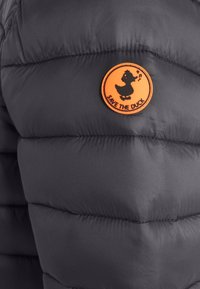 Dunkelgraue Steppjacke mit gesteppten Abschnitten, ausgestattet mit einem runden orangefarbenen Patch mit einem Entenlogo und dem Schriftzug "SAVE THE DUCK" in Schwarz.