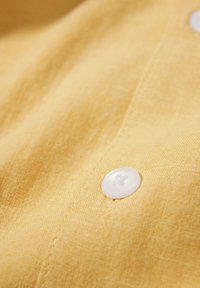 Tessuto di lino giallo con una consistenza liscia, caratterizzato da un grande bottone bianco con quattro fori, posizionato lungo il bordo del tessuto.