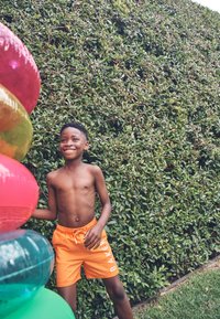 Osmijehni dječak u narančastim kratkim hlačama stoji vani pored naslaganih šarenih napuhavajućih prstenova za bazen, dok se u pozadini vidi zeleni živica od lišća.