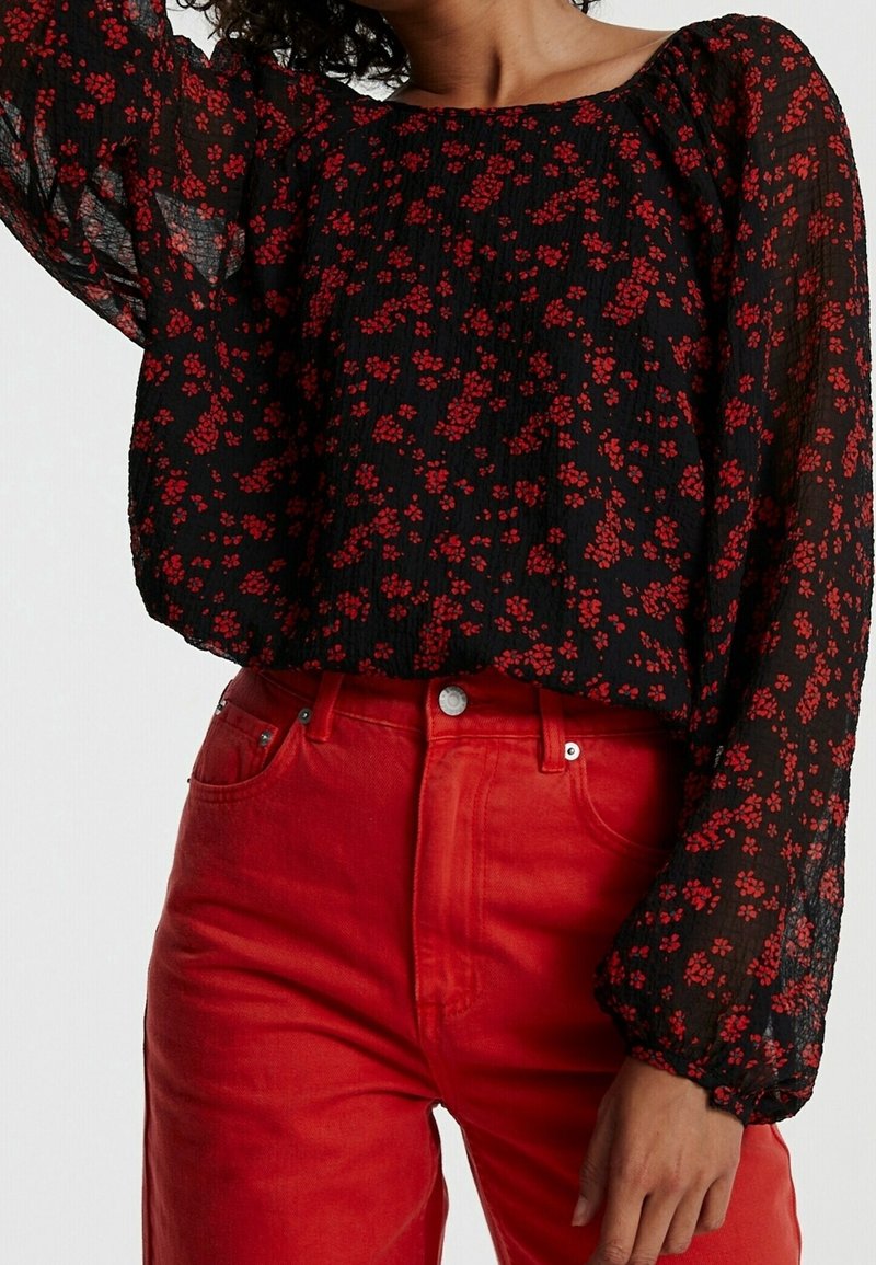 Femme portant un chemisier noir transparent à manches longues avec de petites fleurs rouges et un pantalon taille haute rouge vif.