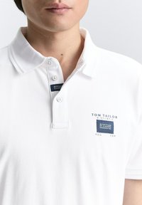 Valge puuvillane polo särk, millel on klassikaline krae, kolme nupuklappi ja sinine ristkülikukujuline logopatch rinnas.