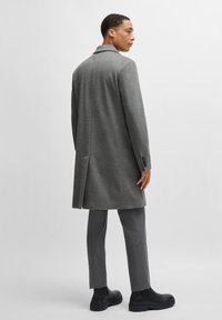 Grau melierter Wollmischungs-Mantel mit taillierter Passform, ausgestattet mit einer Schlitze am Rücken und schwarzen Knopfdetails, kombiniert mit grauen Hosen und schwarzen Stiefeln.