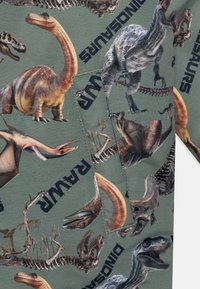 Zaļš audums ar dažādiem dinozauru attēliem un fosiliju dizainiem, ar vārdu "DINOZAUR" izcili redzamā melnā krāsā.