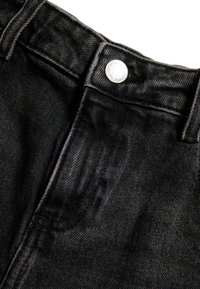 Jeans de mezclilla negra con cierre de botón, una textura sutil y un acabado suave con un ligero brillo. Close-up del área de la cinturilla.