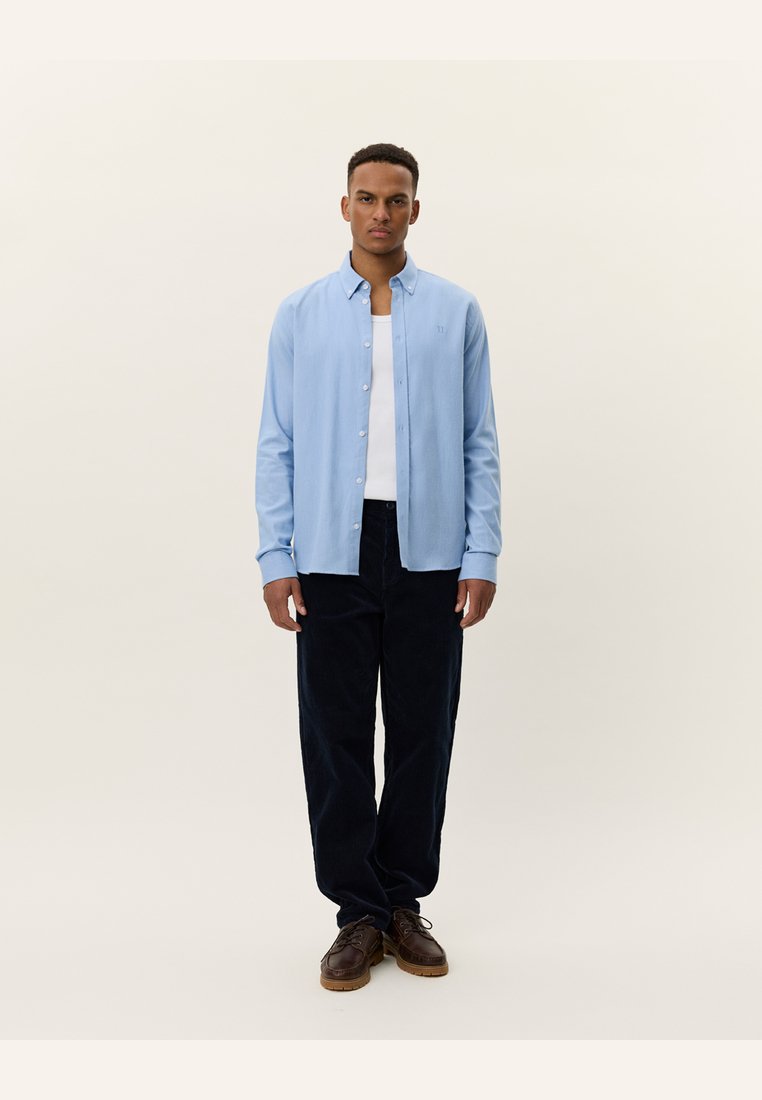 Light blue button-up shirt with long sleeves, worn over a white undershirt, paired with dark corduroy pants and brown shoes.