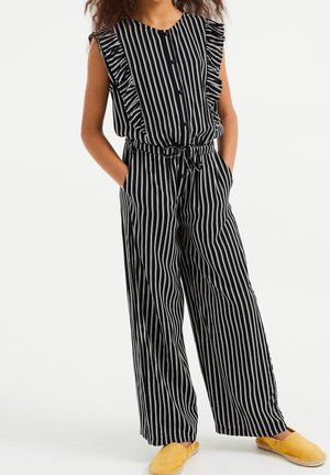 Musta-valkoraidallinen haalari röyhelöhihoilla, etunapeilla, joustavalla vyötäröllä ja leveillä lahkeilla, yhdistettynä keltaisiin liukusäädettäviin kenkiin.