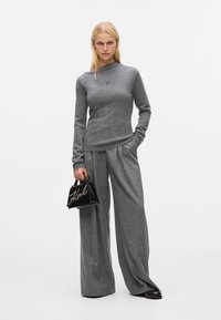 Gray knit sweater with a high neck, paired with wide-leg gray trousers. A small black handbag with a decorative pearl design is held in hand.