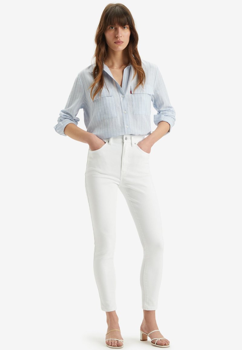 Camicia azzurra a righe con bottoni e due tasche sul petto, abbinata a jeans bianchi attillati e sandali con tacco beige. Sfondo semplice.