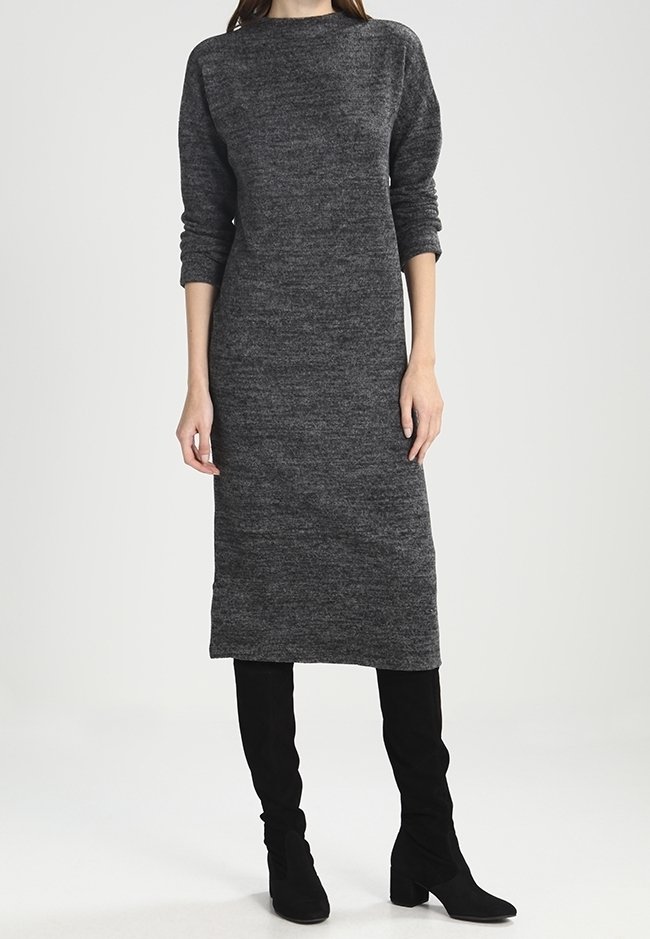 Femme portant une robe en maille gris foncé à manches longues, longueur genou, associée à des bottes noires hautes à talons, debout devant un fond uni.