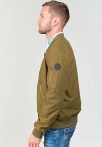 Groene bomberjack met een geribbelde onderkant, voorzien van gestructureerde stof, zijzakken en een patch op de linkermouw. Draagt een patroonhemd eronder.