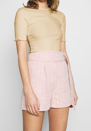 Woman wearing beige short-sleeve ribbed top and high-waisted pink textured shorts with a side tie belt, standing against a plain background.