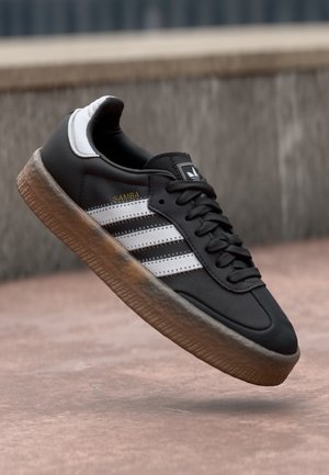 Black Adidas Samba sneaker with white stripes and gum sole, viewed mid-air above a concrete surface and wall background.