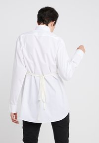 Vit långärmad skjorta med en rundad nertill och detalj med knytning bak. Slät bomullstyg med klassisk krage och knäppning vid ärmsluten.