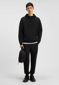 Schwarzer Kapuzenpullover mit strukturiertem Logo, kombiniert mit schwarzen Jogginghosen. Accessoires umfassen eine kleine schwarze Tasche. Schwarze Sportschuhe.