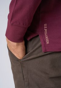 Maglietta rossa a maniche lunghe in tessuto texturizzato, con un logo ricamato in beige vicino all'orlo. Indossata con pantaloni marrone scuro e mano in tasca.