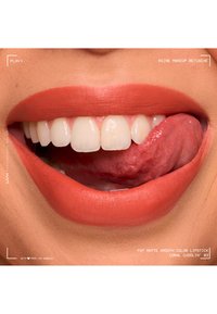 Close-up of smiling lips with coral matte lipstick and visible white teeth as the tongue touches the upper teeth.