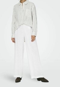 Camicia di lino con bottoni e righe verticali, una tasca sul petto, abbinata a pantaloni bianchi a gamba larga. Le calzature presentano un tacco basso e una superficie texture.