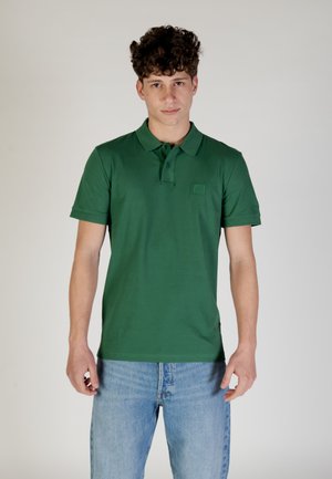 Polo verde con colletto strutturato, maniche corte e un piccolo logo quadrato sul petto, abbinato a jeans blu scoloriti.