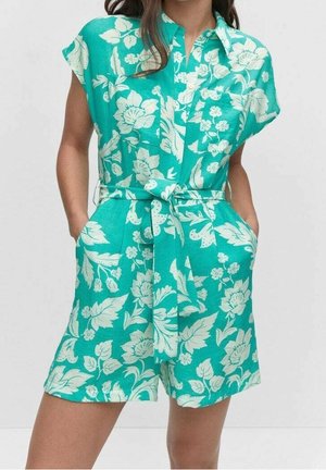 Kvinde iført en teal kortærmet romper med hvid blomsterprint og bundet bælte i taljen, hænder i lommerne, stående mod en ensfarvet baggrund.