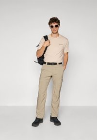 Ljusbeige t-shirt med korta ärmar och logotyp, kombinerad med ljusa khakifärgade cargobyxor och svarta vandringsskor. Accessoarer inkluderar svart ryggsäck och solglasögon.