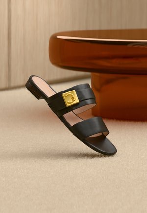 Sandale en cuir noir avec deux brides, détail de boucle carrée dorée, présentée sur un tapis beige et un pied de table rond marron.