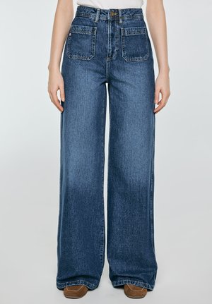 Modré džíny s vysokým pasem a širokými nohavicemi, s předními našitými kapsami a knoflíkovým zapínáním, nošené s hnědými botami a bílým topem.