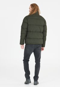 Puffjakke i mørkegrøn, med kviltet design og høj krave. Båret med slanke sorte jeans og ankelstøvler. Bagsidevisning vist.