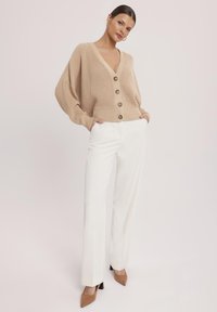 Beżowy sweter z dzianiny z dekoltem w serek i guzikami z przodu, zestawiony z białymi spodniami o szerokich nogawkach i brązowymi szpilkami w szpic. Prosty, teksturowany design.