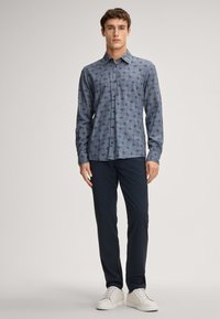 Donkerblauw overhemd met een golvend geometrisch patroon, lange mouwen en een puntkraag. Gecombineerd met donkere broeken en witte sneakers.