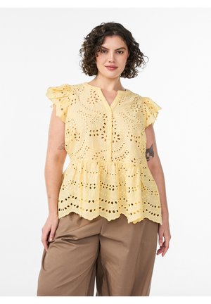 Kobieta z kręconymi włosami, ubrana w żółtą bezrękawnikową bluzkę z ażurowym wzorem i falbankami na rękawach oraz brązowe spodnie z szerokimi nogawkami, stojąca na białym tle.