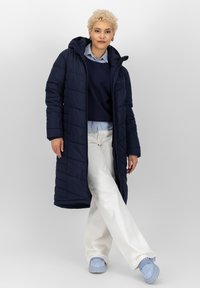 Persoon staat met één been gekruist, gekleed in een marineblauwe lange gewatteerde jas, een blauwe trui, een lichtblauw shirt, witte broek en lichtblauwe schoenen.