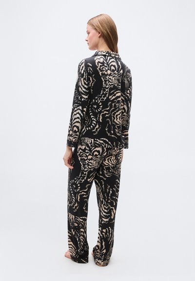 Γυναίκα όρθια με γυμνά πόδια, στραμμένη προς τα αριστερά, φοράει μαύρη, άνετη πυτζάμα με μπεζ αφηρημένα στροβιλιστά σχέδια.