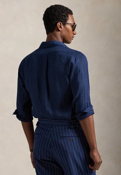 Hombre con camisa de manga larga azul oscuro con puños remangados y pantalones azul marino con rayas finas, de espaldas sobre un fondo liso.