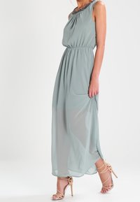 Bezramínková maxi šaty ve světle zeleném chiffon s nařaseným pasem a zdobeným výstřihem. V kombinaci s béžovými sandály na vysokém podpatku.