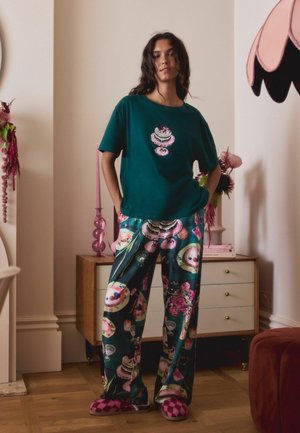 Tricou verde din bumbac cu un grafic de tort; pantaloni de pijama cu un model în tonuri închise, prezentând deserturi; papuci roz în carouri. Mobilier din lemn în fundal.