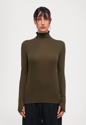 Eine Frau mit dunklem Haar trägt einen olivegrünen, taillierten Rollkragenpullover mit Rüschensaum und schwarze weit geschnittene Hosen und posiert vor einem weißen Hintergrund.