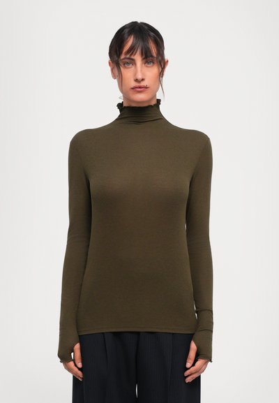 Una donna con i capelli scuri indossa un maglione aderente a collo alto verde oliva con colletto arricciato e pantaloni neri a gamba larga, posando su uno sfondo bianco.