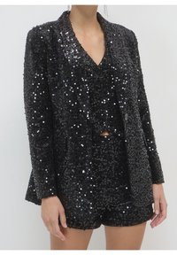 Blazer noir à sequins et short assorti portés sur un haut noir à sequins sur une personne debout devant un fond uni.