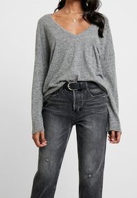 Graues Langarm-V-Ausschnitt-Oberteil mit lockerer Passform, ausgestattet mit einer Fronttasche, kombiniert mit hoch taillierten schwarzen Jeans mit abgenutzten Details.