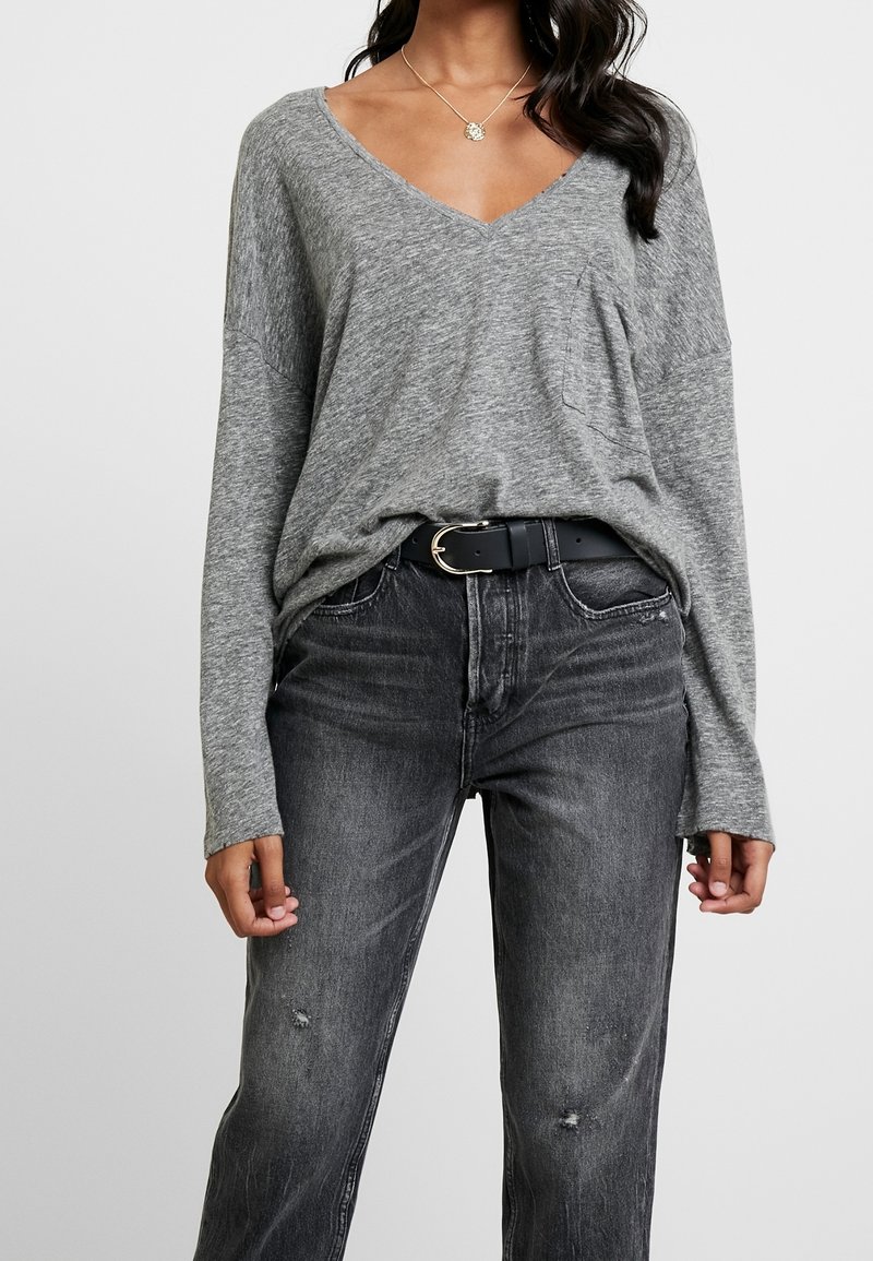 Graues Langarm-V-Ausschnitt-Oberteil mit lockerer Passform, ausgestattet mit einer Fronttasche, kombiniert mit hoch taillierten schwarzen Jeans mit abgenutzten Details.