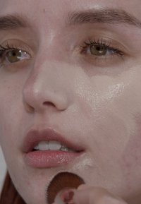 Nahaufnahme eines Gesichts, das sich mit einem Pinsel schminkt. Die Haut hat ein feuchtes Finish, leichte Unreinheiten sind sichtbar und die Lippen sind in einem natürlichen Rosa.