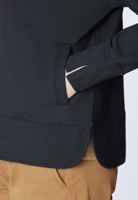 Pullover nero dalla superficie liscia, con una fessura laterale e una tasca nascosta, abbinato a pantaloni marrone chiaro.