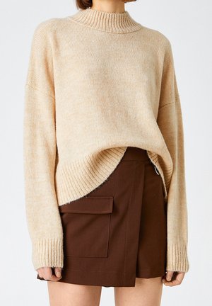 Beige knitted sweater with a high ribbed neckline and dropped shoulders, paired with a brown wrap skirt featuring side pockets and a metal accent.