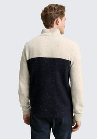Mann in einem gesprenkelten schwarzen und beigen Langarmstrickpullover mit hohem Kragen, der von einem schlichten Hintergrund abgewandt ist.