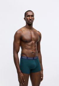 Zöld boxer alsónadrág sötétkék derékrésszel, amelyen a "PUMA" logó látható, testhezálló fazonban, bemutatva egy izmos férfi modellen, egyszínű háttér előtt.