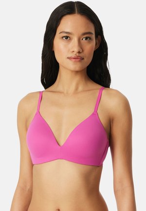 Mujer con el pelo largo y oscuro, vistiendo un sostén inalámbrico rosa brillante con tirantes delgados ajustables, sobre un fondo liso.