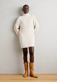 Fehér, bordás kötött pulóverruha magas gallérral és oversized ujjakkal. Barna bőr cowboy csizmával viselve, hegyes orral.