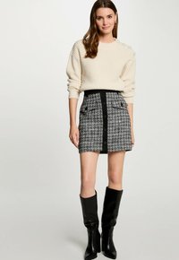 Beige gestrickter Pullover mit seitlichen Knöpfen, kombiniert mit einem schwarz-weißen Tweed-Minirock mit vorderen Knöpfen und seitlichen Taschen. Schwarze Stiefel bis zum Knie.